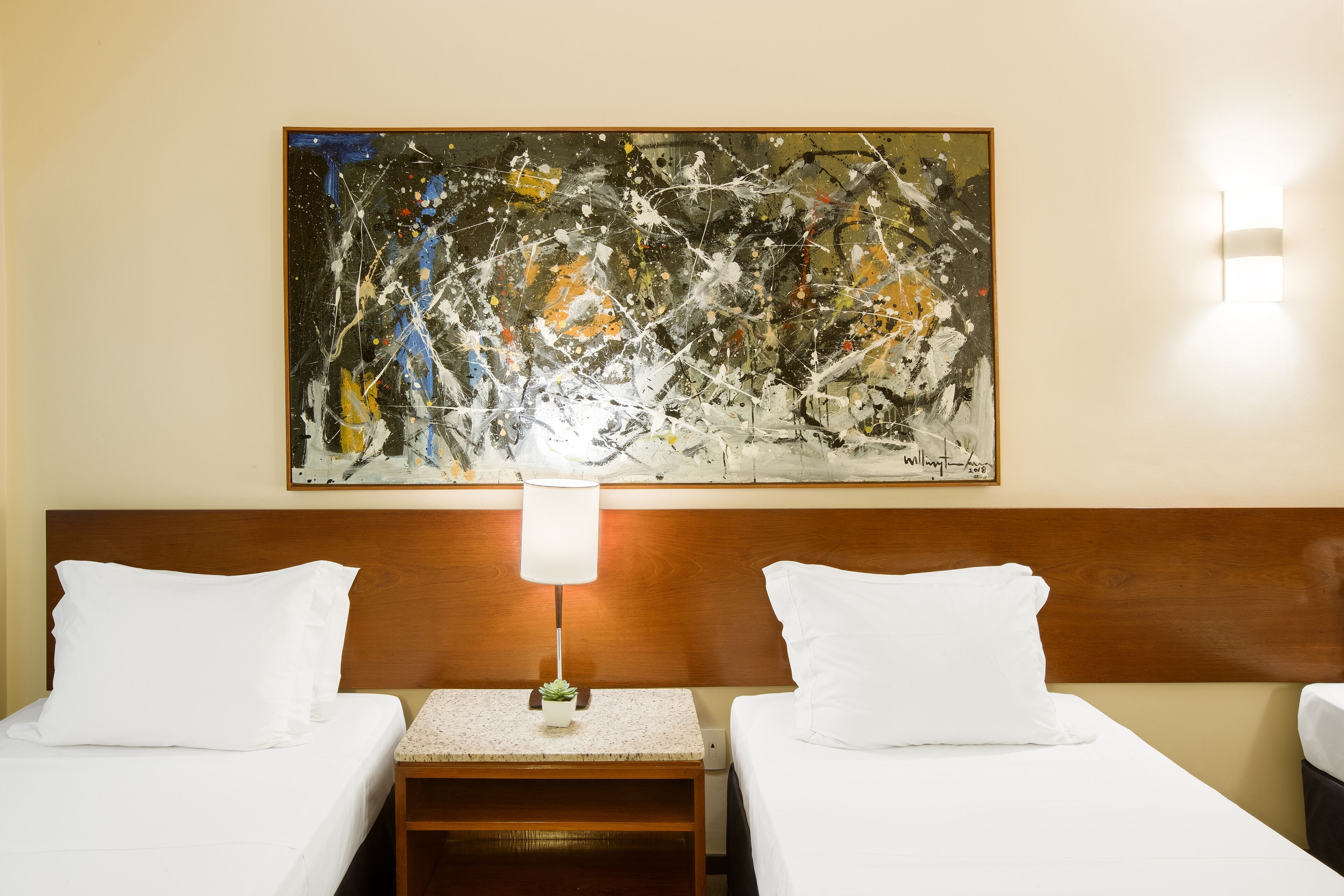 Habitación de hotel con dos camas, cabecero de madera, un cuadro abstracto en la pared, una mesita con lámpara y una mesita de noche.