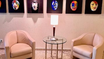 Um quarto com duas poltronas bege, uma mesa de vidro com abajur, e uma parede com seis máscaras coloridas.