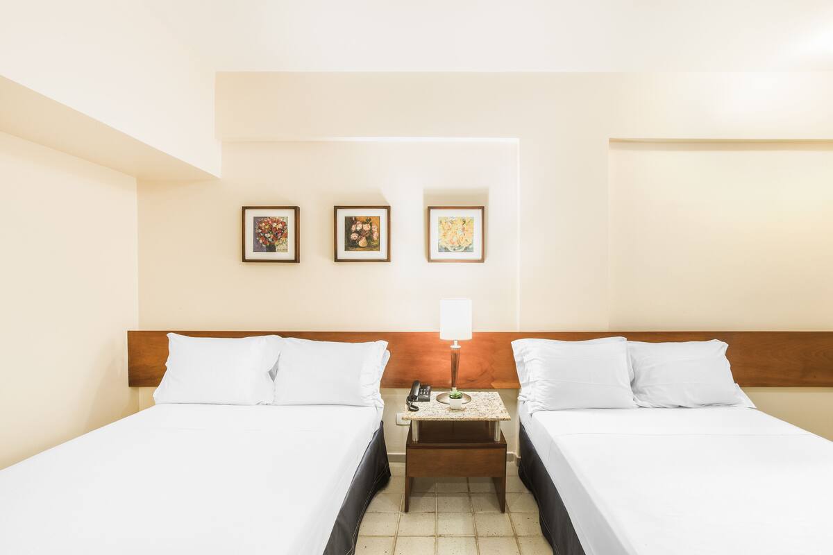 A hotel room with two beds, a nightstand with a lamp, and three framed pictures on the wall.