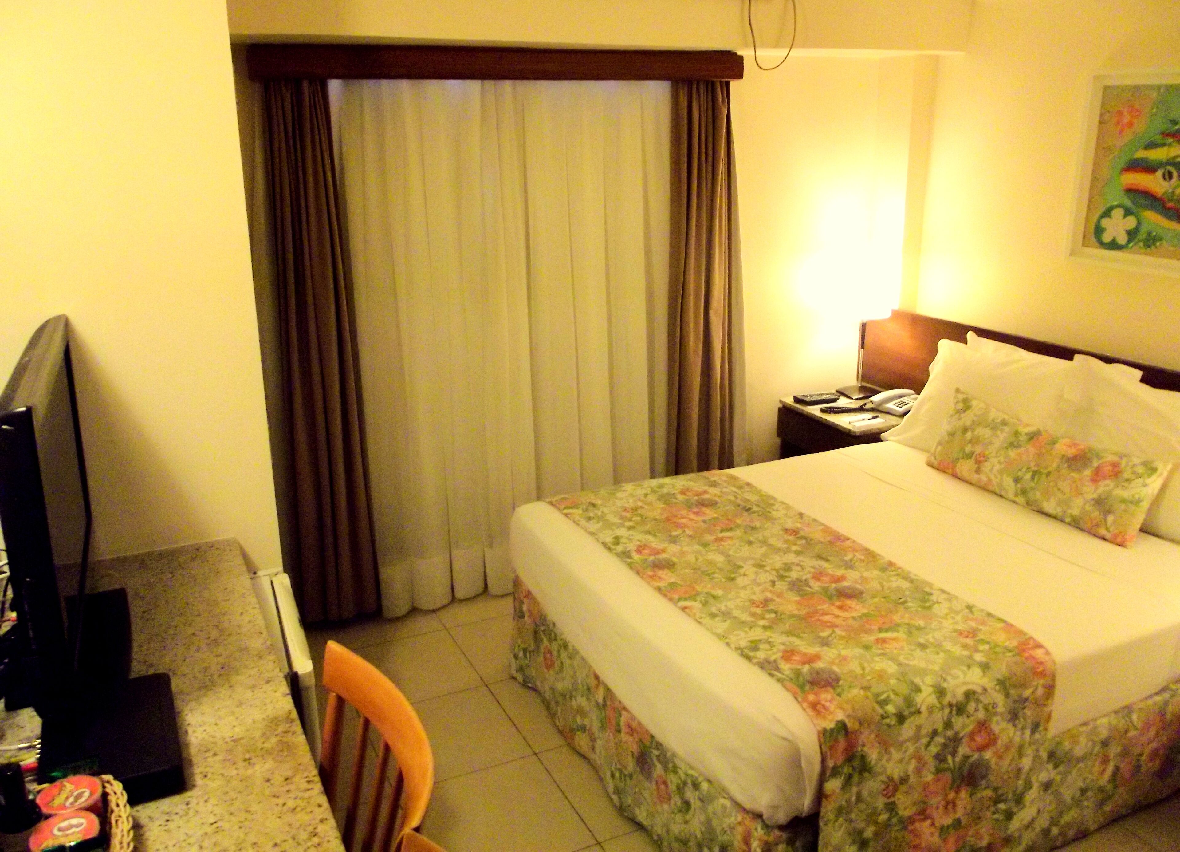 Habitación de hotel con cama, escritorio y silla, televisor y mesita de noche con lámpara.