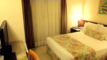 Habitación de hotel con cama, escritorio y silla, televisor y mesita de noche con lámpara.
