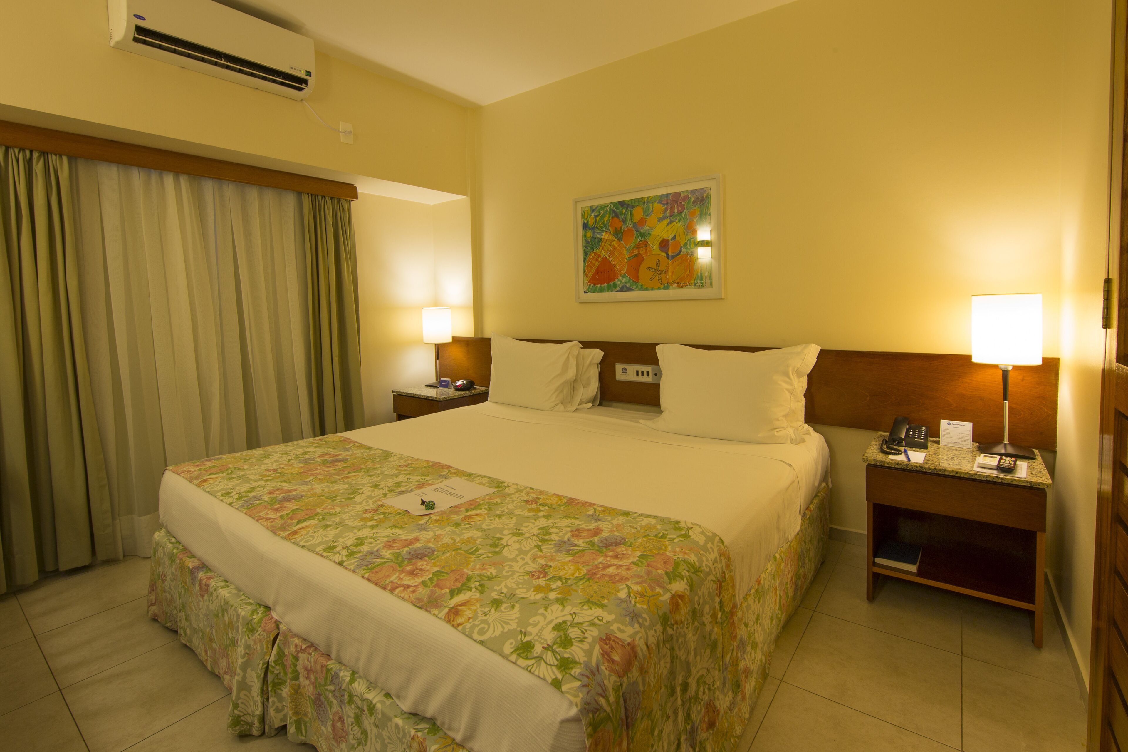 A hotel room with a large bed, two bedside tables with lamps, a painting on the wall, and a window with curtains.