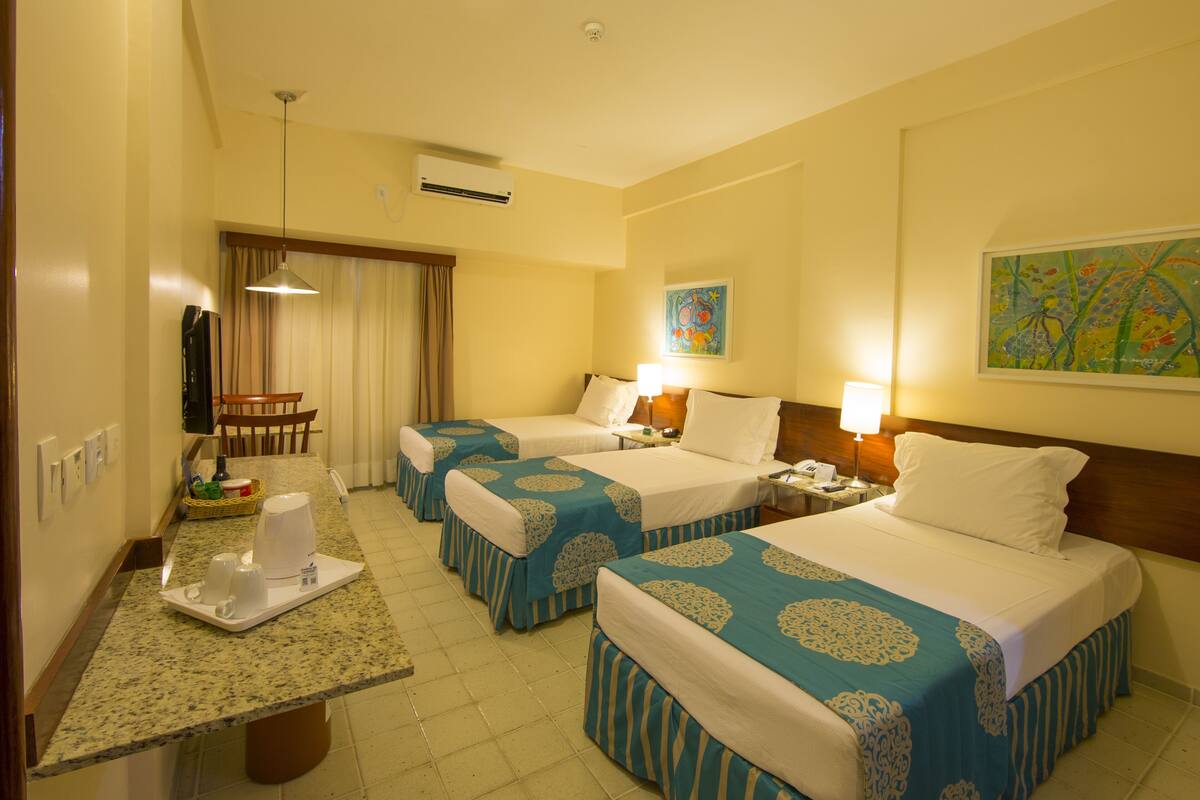A hotel room with three beds, a dining table with a tray of cups, a chair, and a painting on the wall.