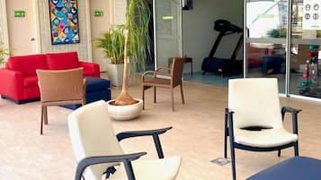 Un área de estar moderna con un sofá rojo, sillones de mimbre y una cinta de correr al fondo.