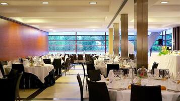 A spacious dining area with multiple tables set for a formal event, large windows providing natural light, and a modern architectural design with high ceilings and columns.