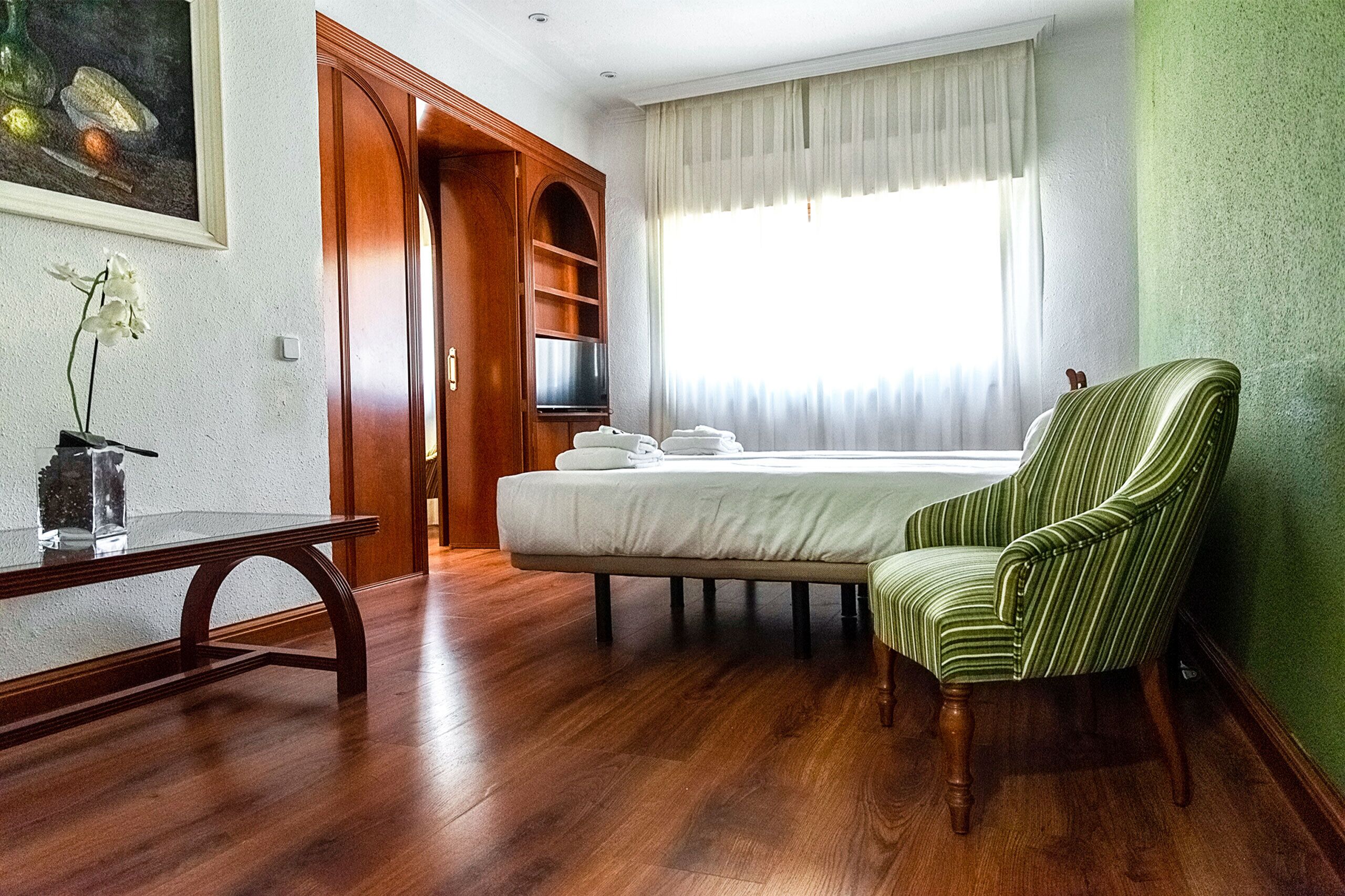 Una habitación de hotel con una cama, un sillón verde a rayas, una mesa de madera y un cuadro en la pared.
