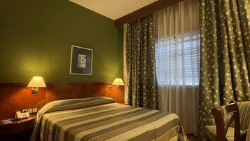 Una habitación de hotel con cama, dos lámparas de noche, mesita de noche, una silla y una ventana con cortinas.