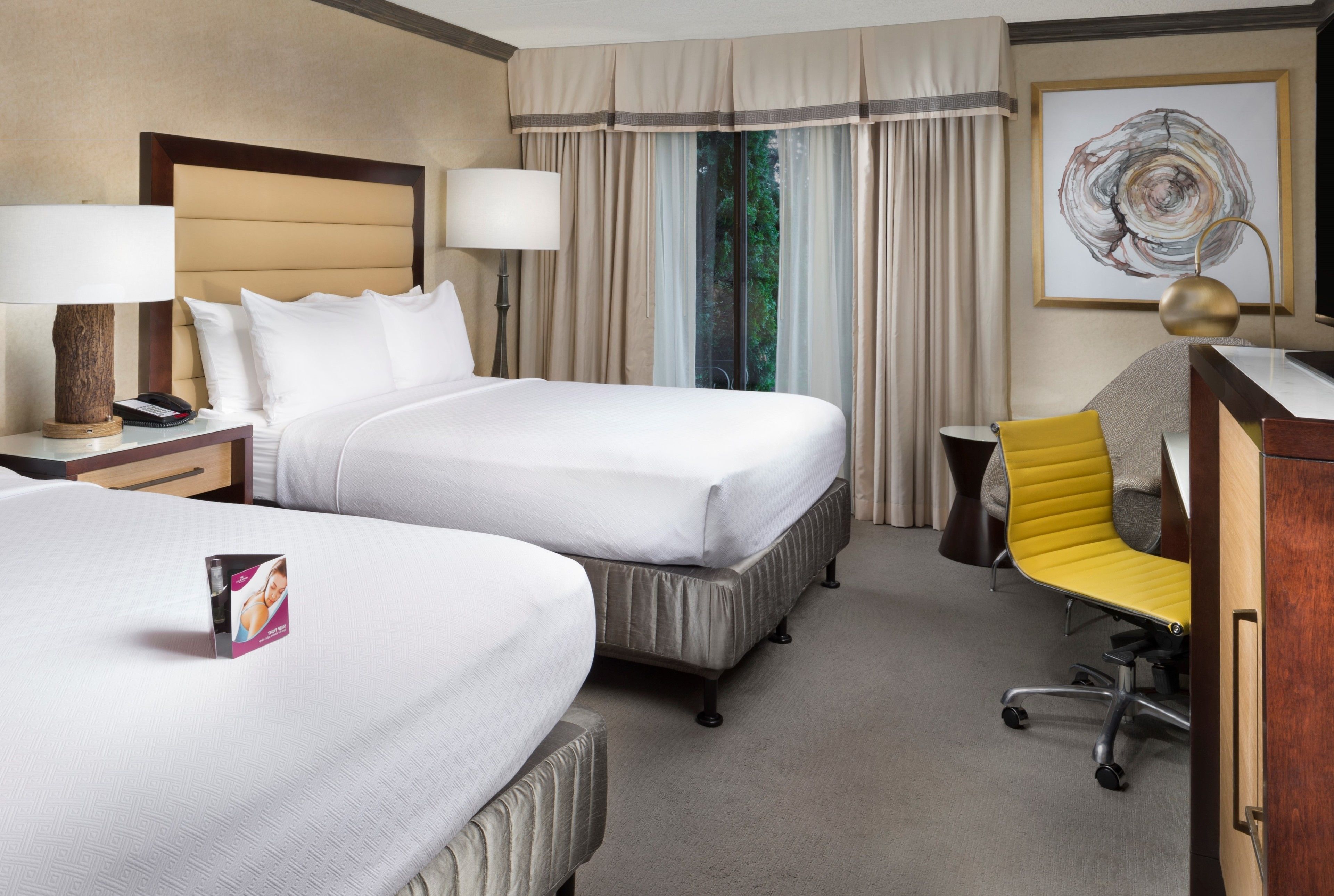A hotel room with two beds, a desk, a chair, and a large window with curtains.