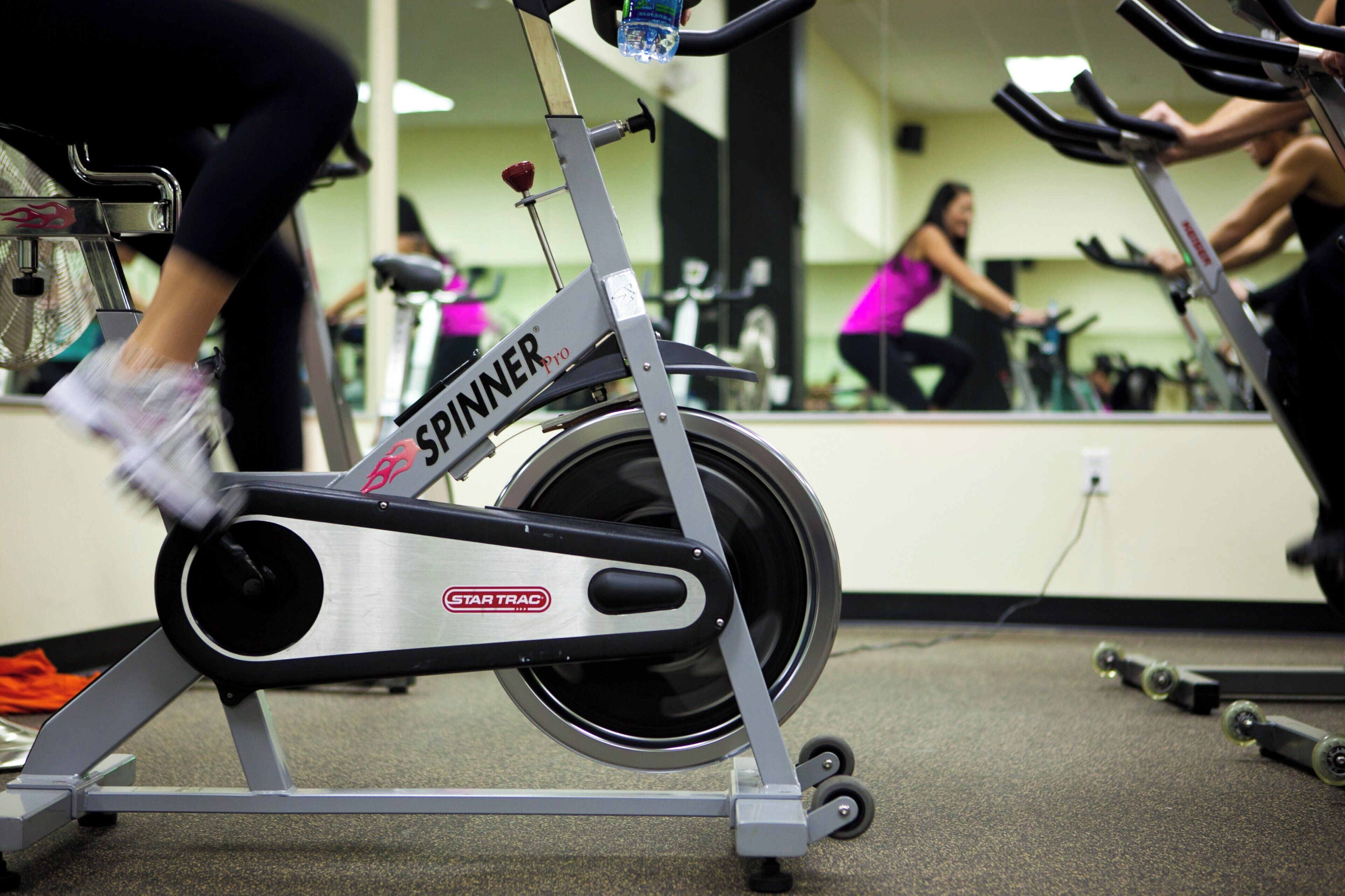 Un vélo d’appartement avec les marques « SPINNER » et « STAR TRAC » bien visibles, ce qui indique qu’il se trouve dans une salle de sport équipée de matériel d’exercice.
