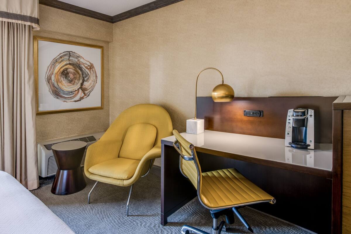 A hotel room with a bed, a yellow armchair, a desk with a coffee maker, and a framed abstract painting.