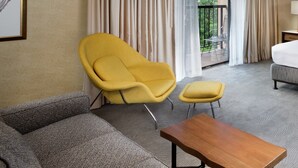 Quarto de hotel com sofá, poltrona amarela, mesa de madeira e varanda com cadeira.