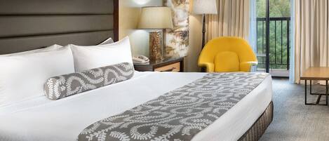 Habitación de hotel con una cama grande, una mesita de noche con lámpara, una silla y vistas al exterior.