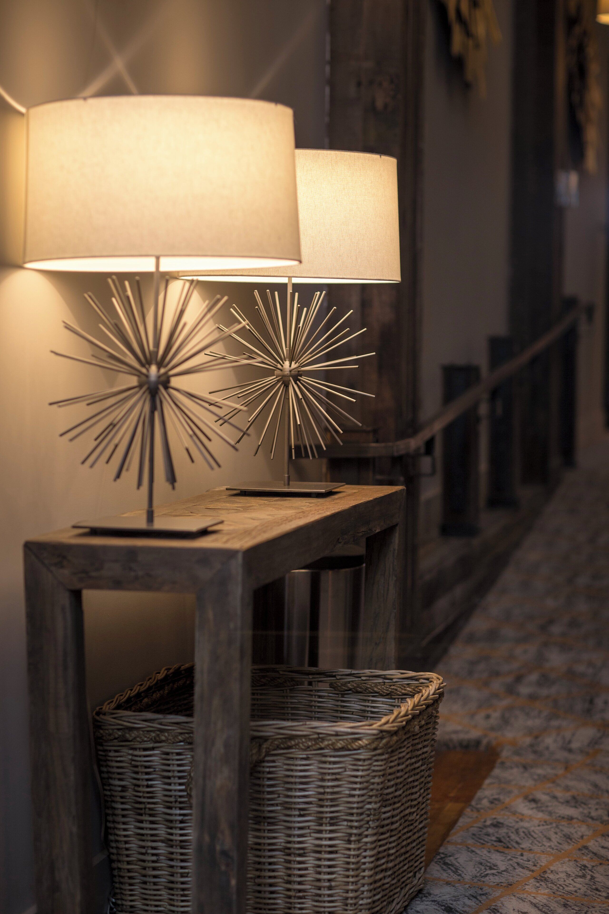 A lamp with a starburst design on a wooden console table, a wicker basket, and a patterned carpet.
