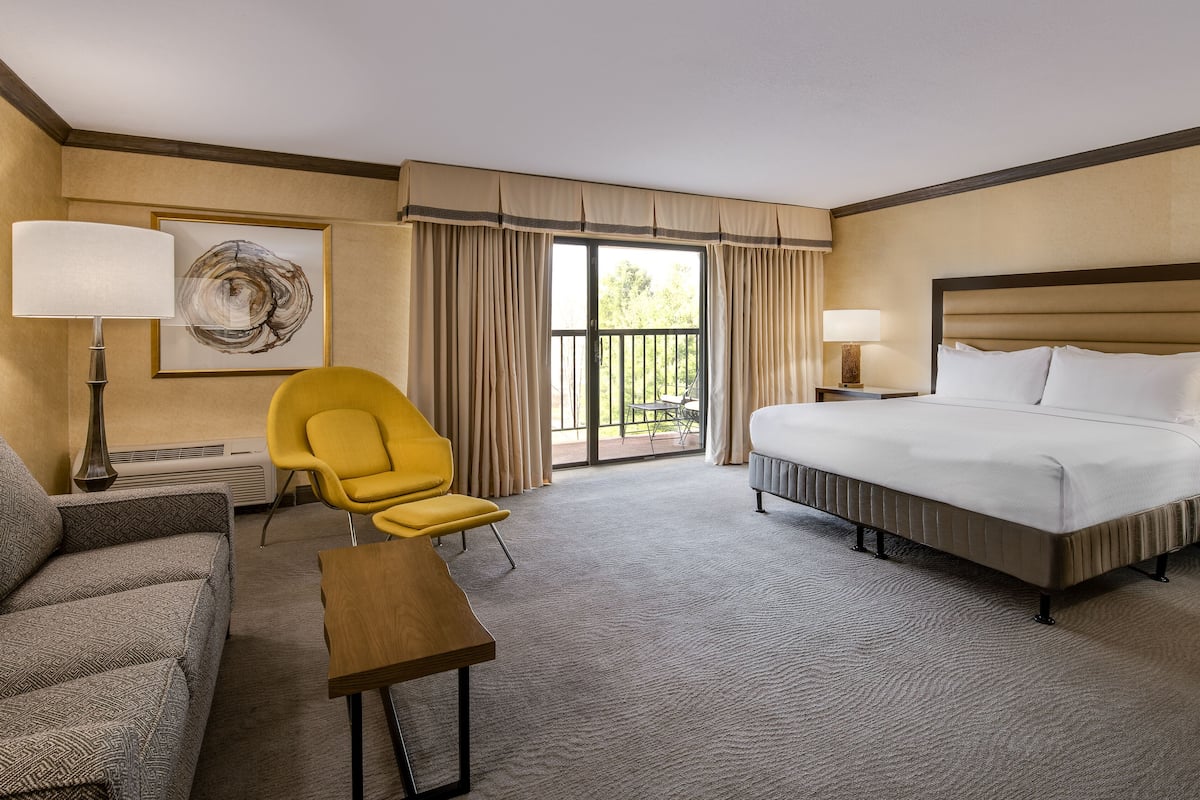 A hotel room with a bed, a chair, a table, a lamp, and a painting on the wall.