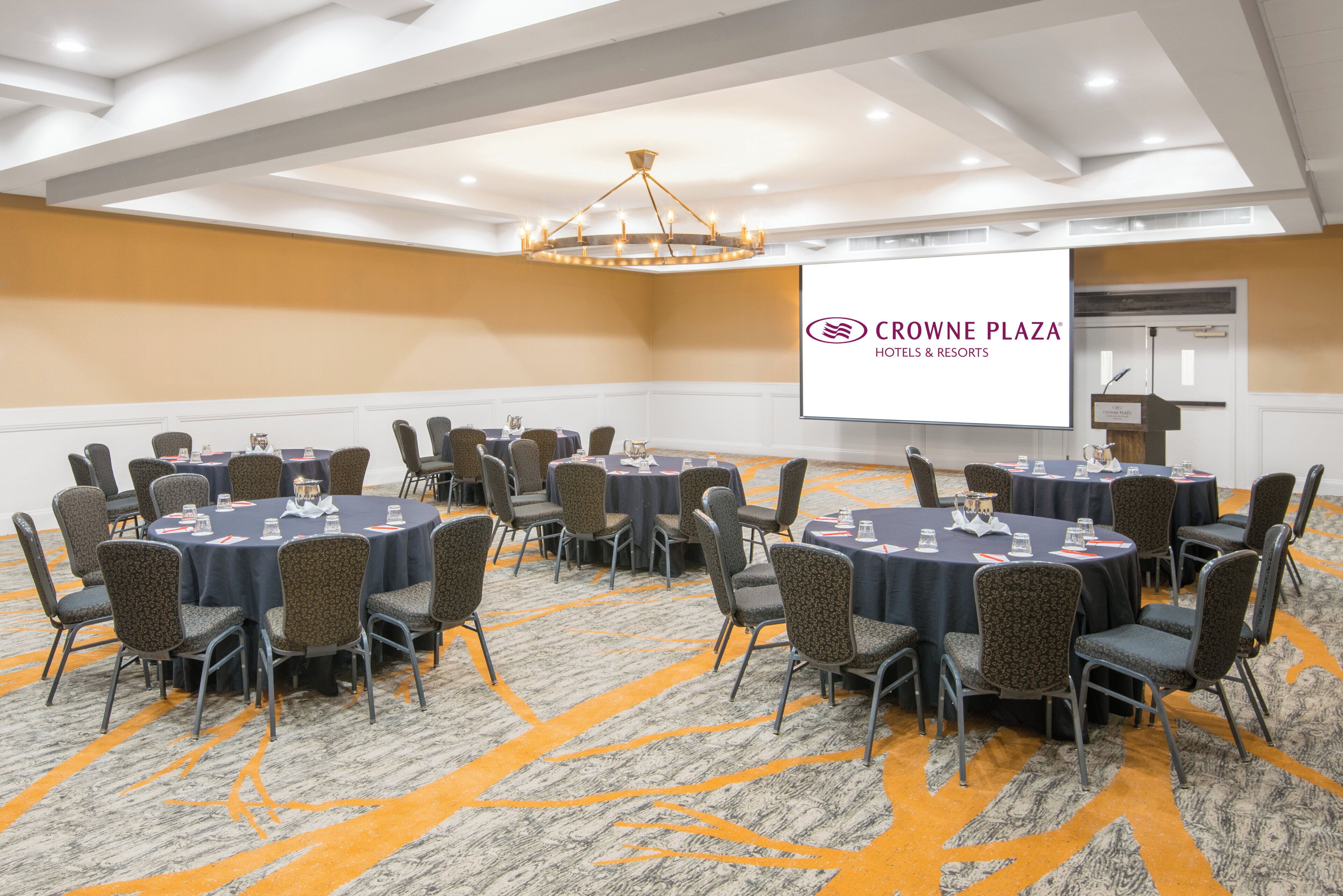 Un salón de conferencias con mesas redondas y sillas, una araña y una pantalla mostrando el logotipo de Crown Plaza Hotels & Resorts.