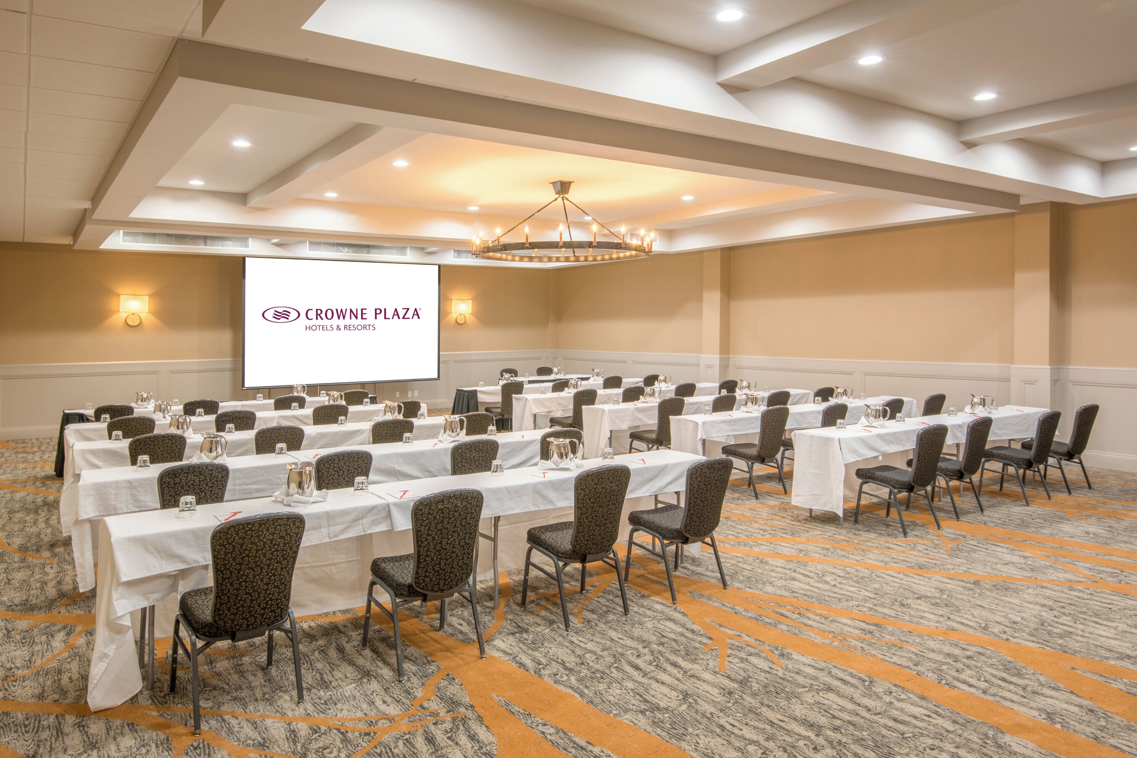A conference room with rows of tables and chairs, a projector screen, and a chandelier.
