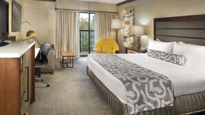 Quarto de hotel com uma cama grande, uma escrivaninha com cadeira, uma poltrona amarela e vista para o exterior pela janela.