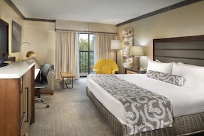 A hotel room with a large bed, a desk with a chair, a yellow armchair, and a view of the outdoors through a window.