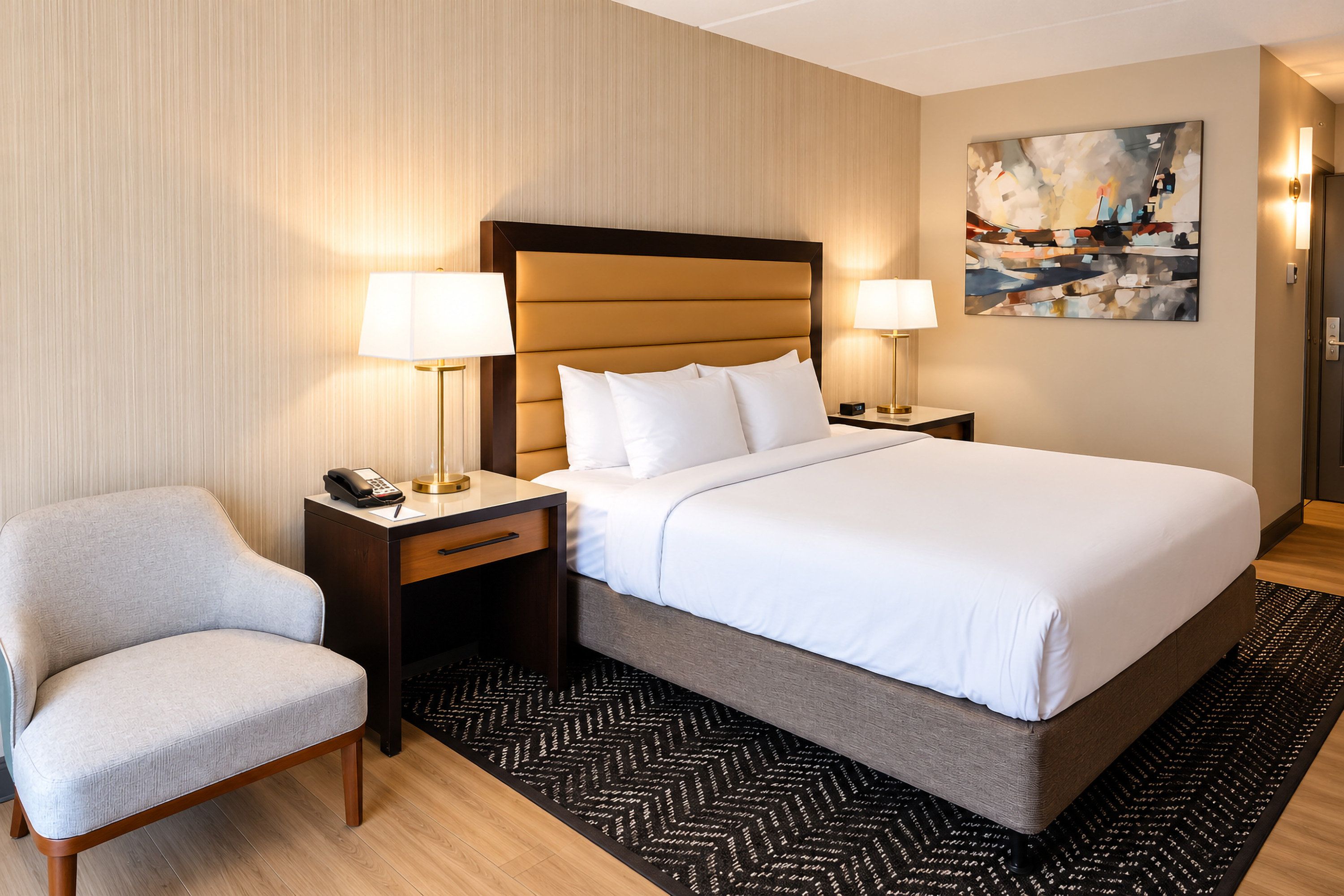 A hotel room with a bed, two bedside tables, a chair, a desk with a telephone, and a painting on the wall.