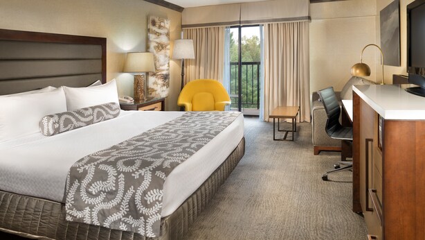 Quarto de hotel com uma cama grande, uma escrivaninha com cadeira, uma poltrona amarela e uma varanda com cortinas.