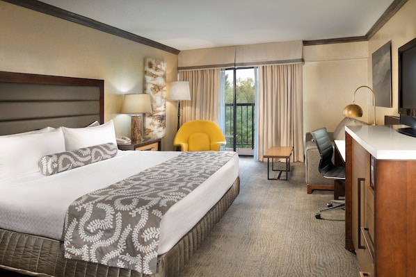 A hotel room with a large bed, a desk with a chair, a yellow armchair, and a balcony with curtains.
