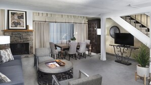 Uma sala de estar com lareira de pedra, uma área de jantar com mesa e cadeiras, um sofá, uma mesa de centro e uma escada com uma TV em um móvel.