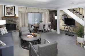 A living room with a stone fireplace, a dining area with a table and chairs, a sofa, a coffee table, and a staircase with a TV on a stand.