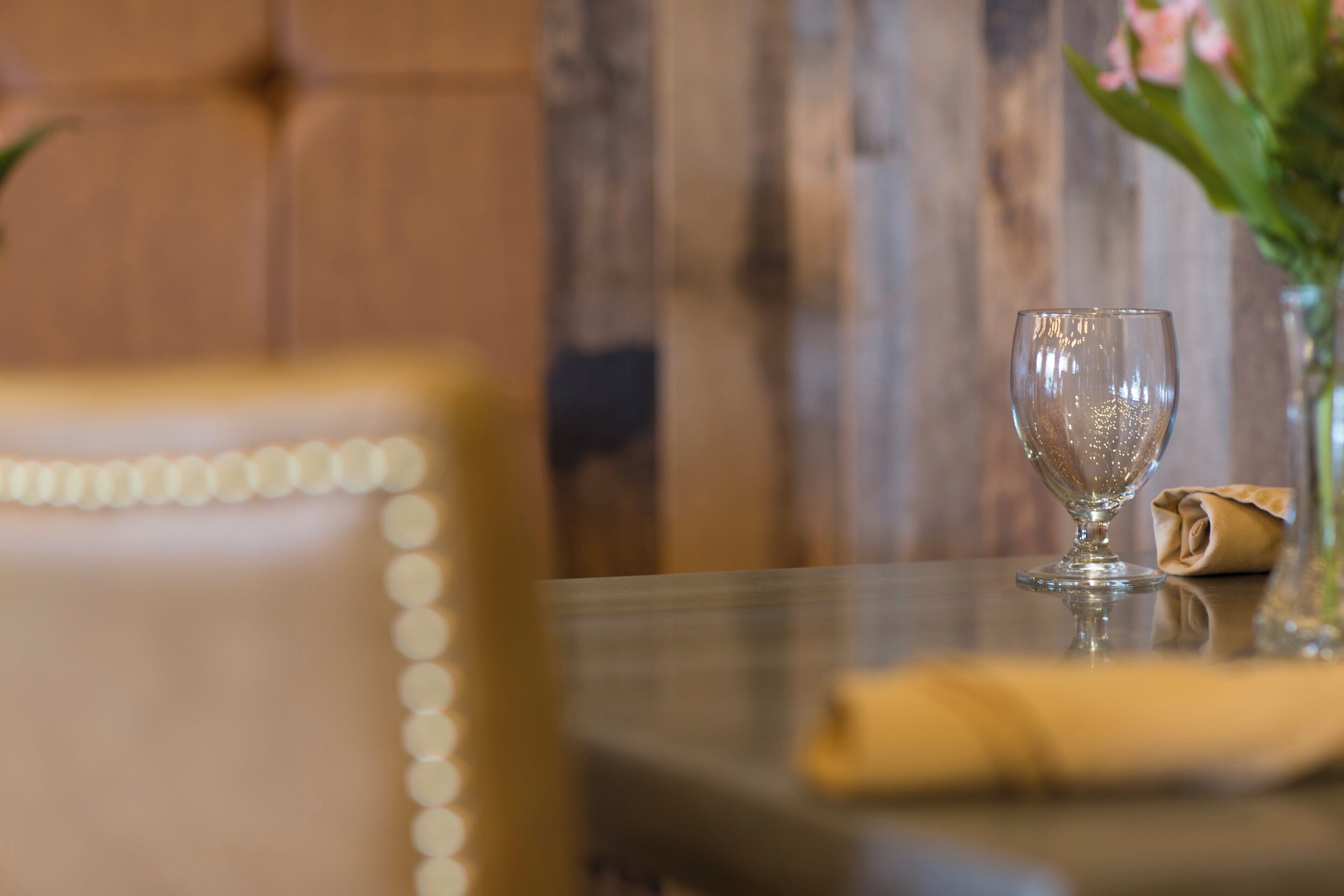 A dining table with a glass and a napkin, a chair with a decorative backrest, and a wooden wall in the background.