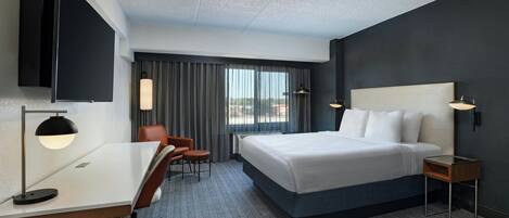 A hotel room with a large bed, a desk with a lamp, a chair, a TV mounted on the wall, and a window with curtains.