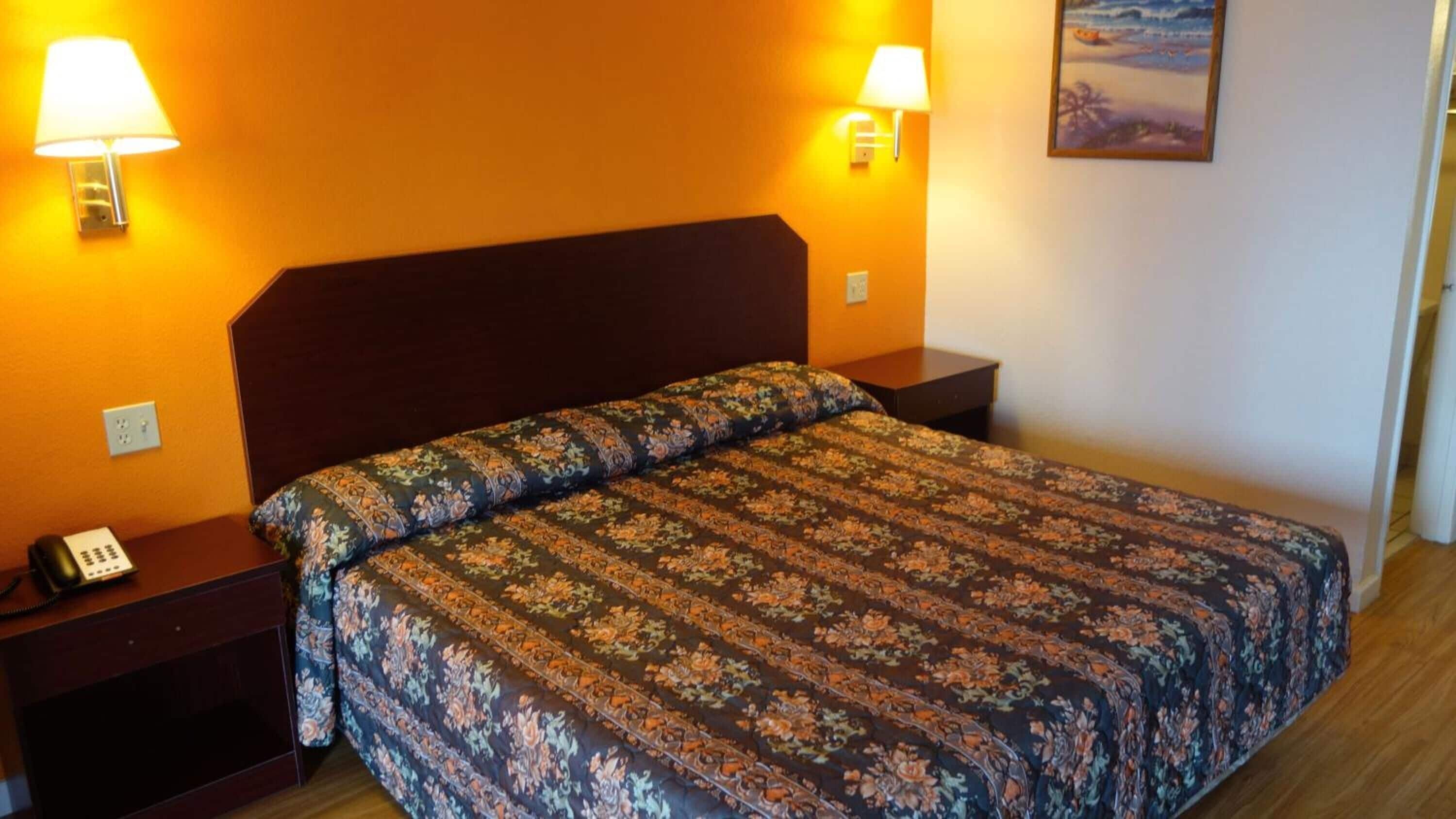 A hotel room with a floral-patterned bedspread, two bedside lamps, a nightstand with a telephone, and a painting on the wall.