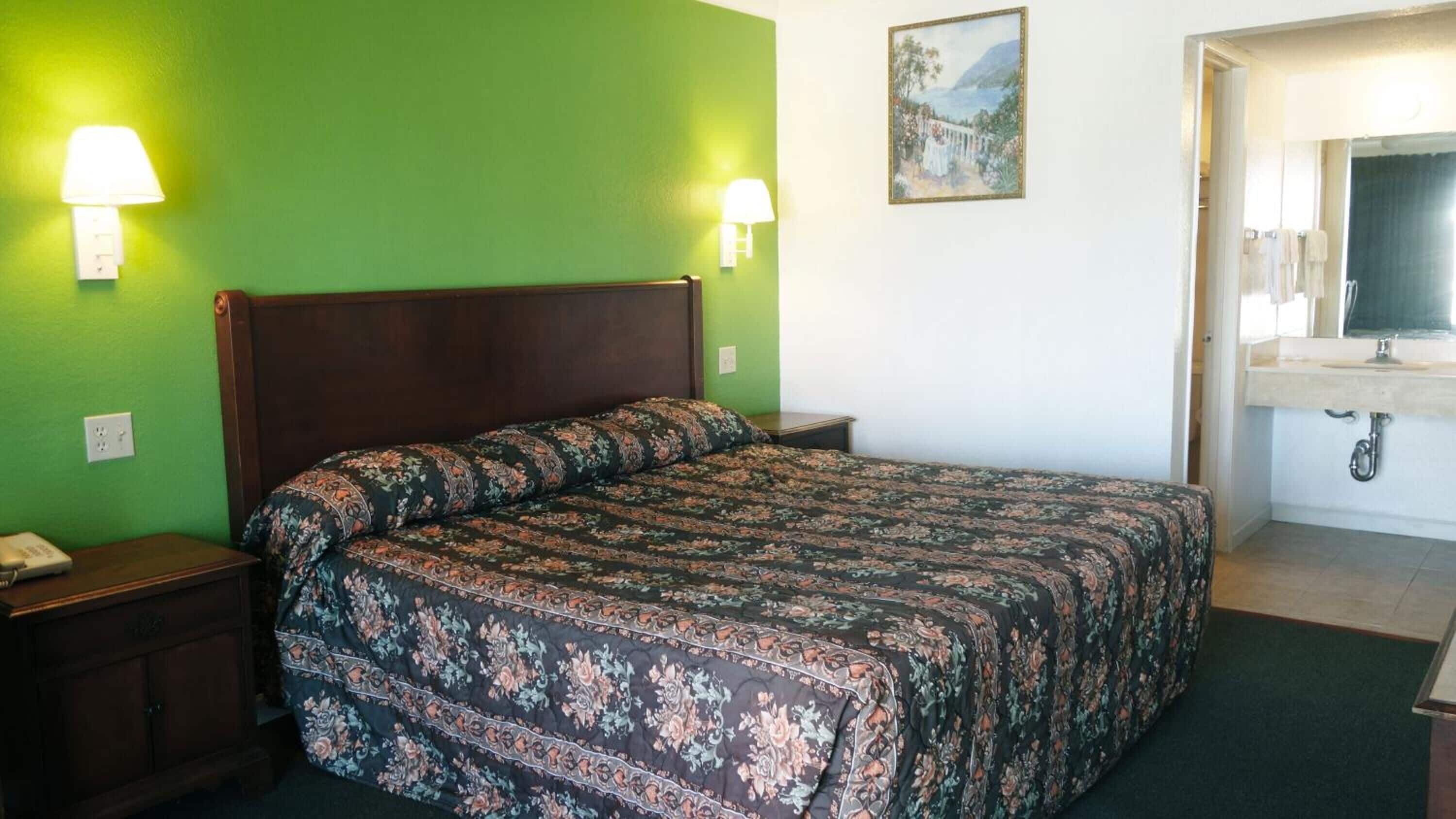 Een hotelkamer met een bed, nachtkastje, lamp, schilderij aan de muur, en een badkamer met een wastafel en spiegel.