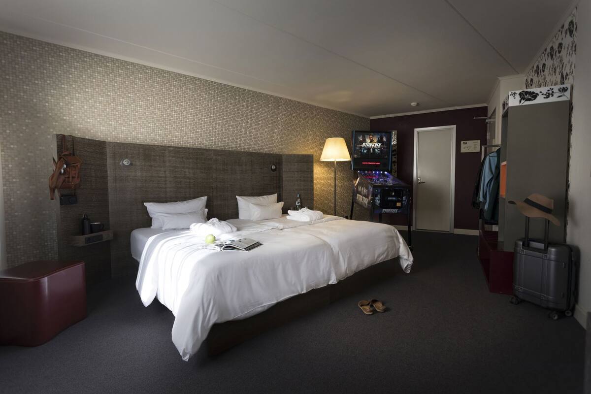 Quarto de hotel com cama grande, mesa de cabeceira, abajur, televisão, guarda-roupa e mala.