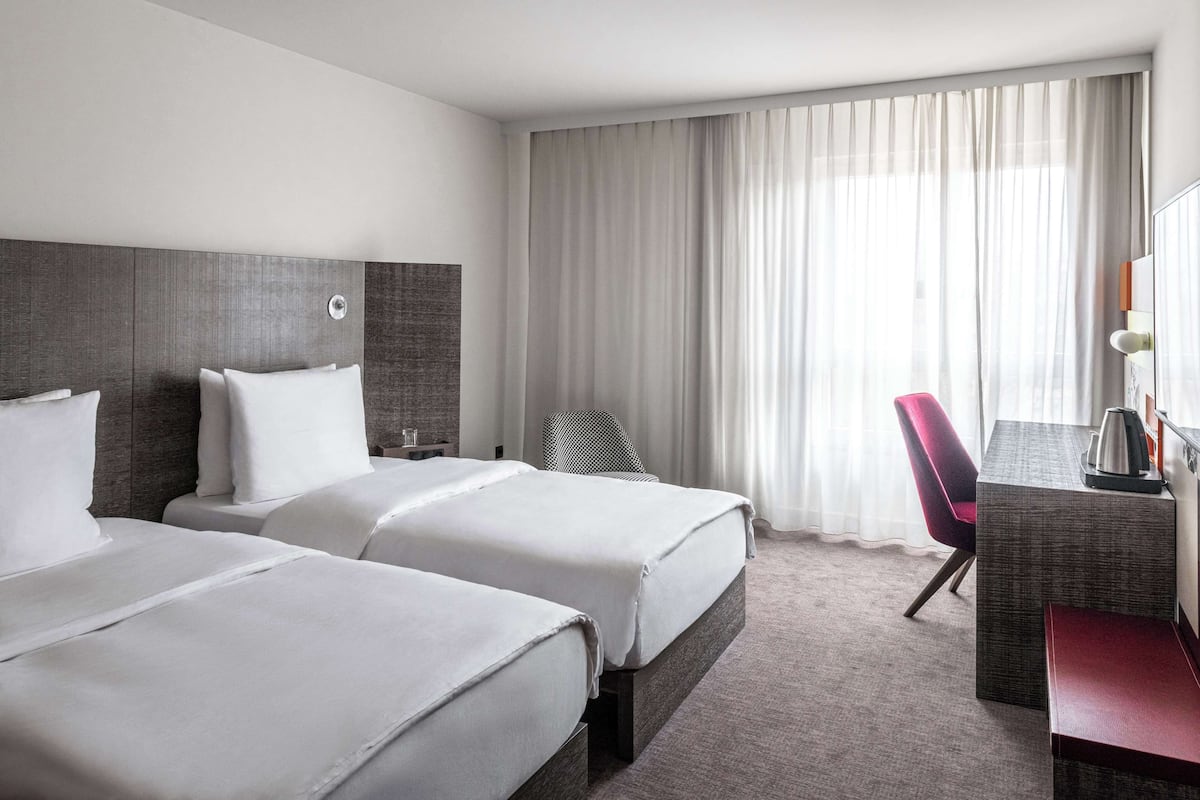 Quarto de hotel com duas camas, uma escrivaninha, uma cadeira e uma janela grande com cortinas.