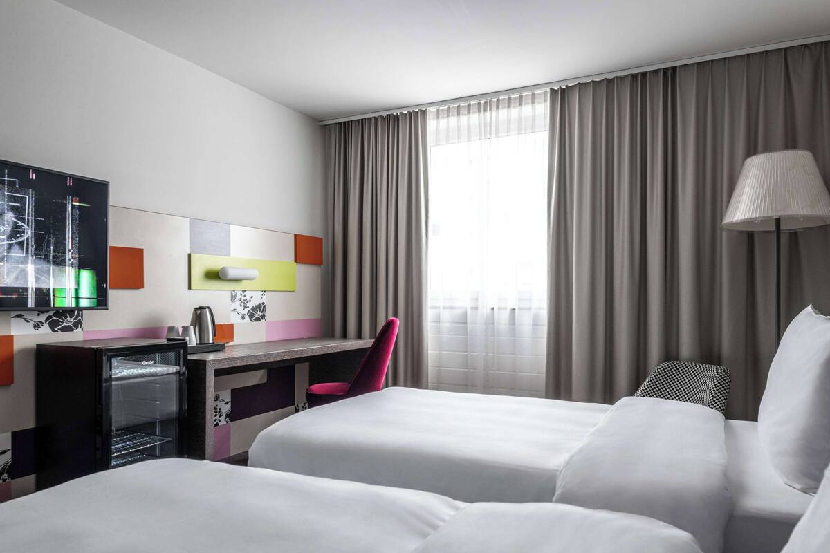 Quarto de hotel com duas camas, uma escrivaninha, uma cadeira, uma televisão e uma janela com cortinas.