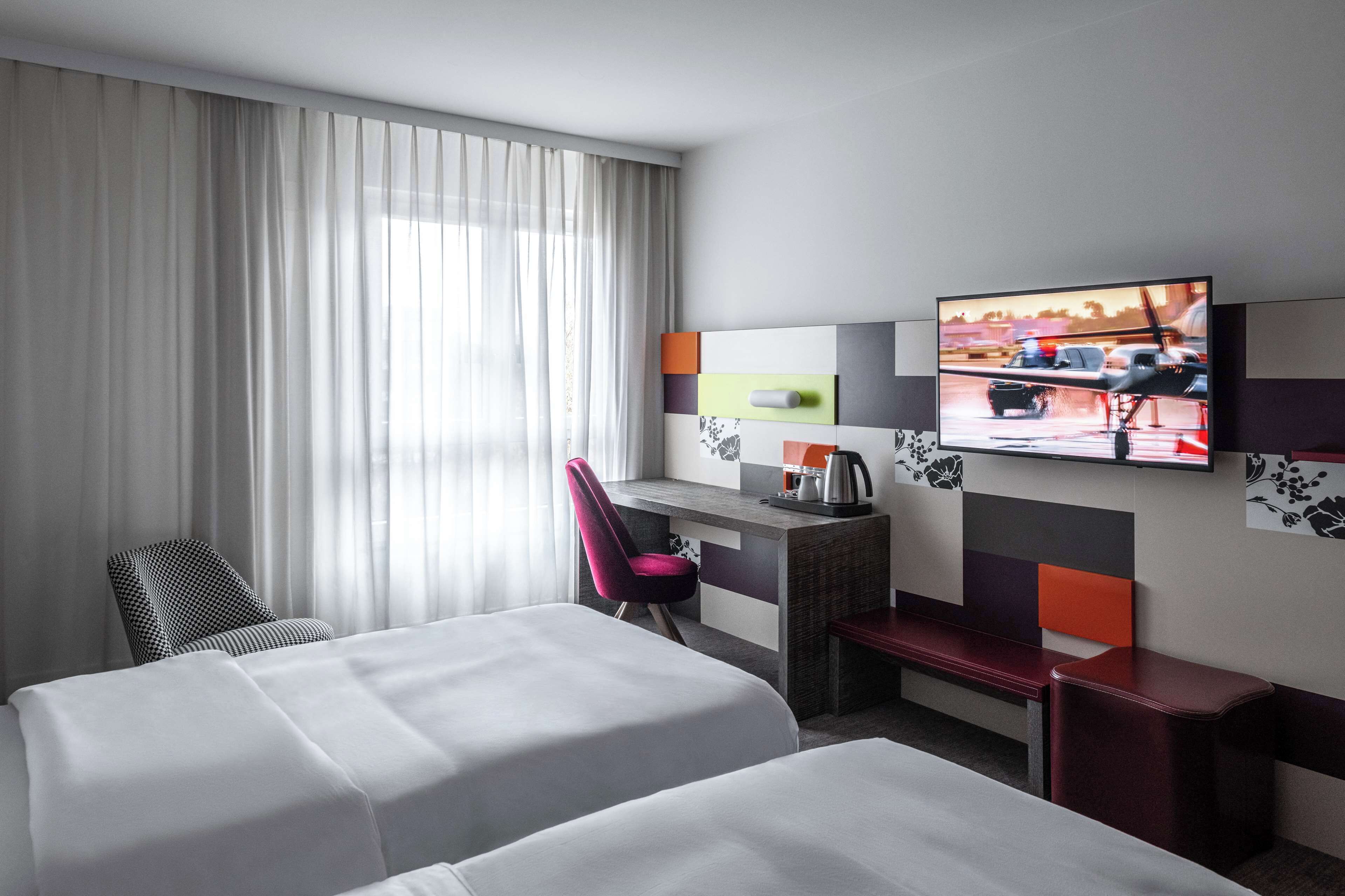 A hotel room with two beds, a desk with a chair, a television on the wall, and a window with curtains.