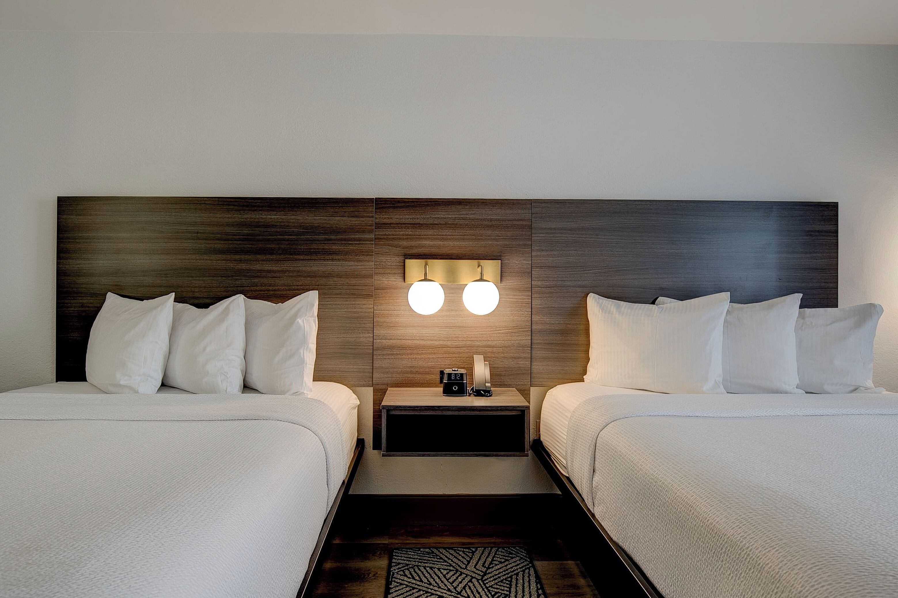 A hotel room with two beds, a nightstand with a lamp, and a wall-mounted light fixture.