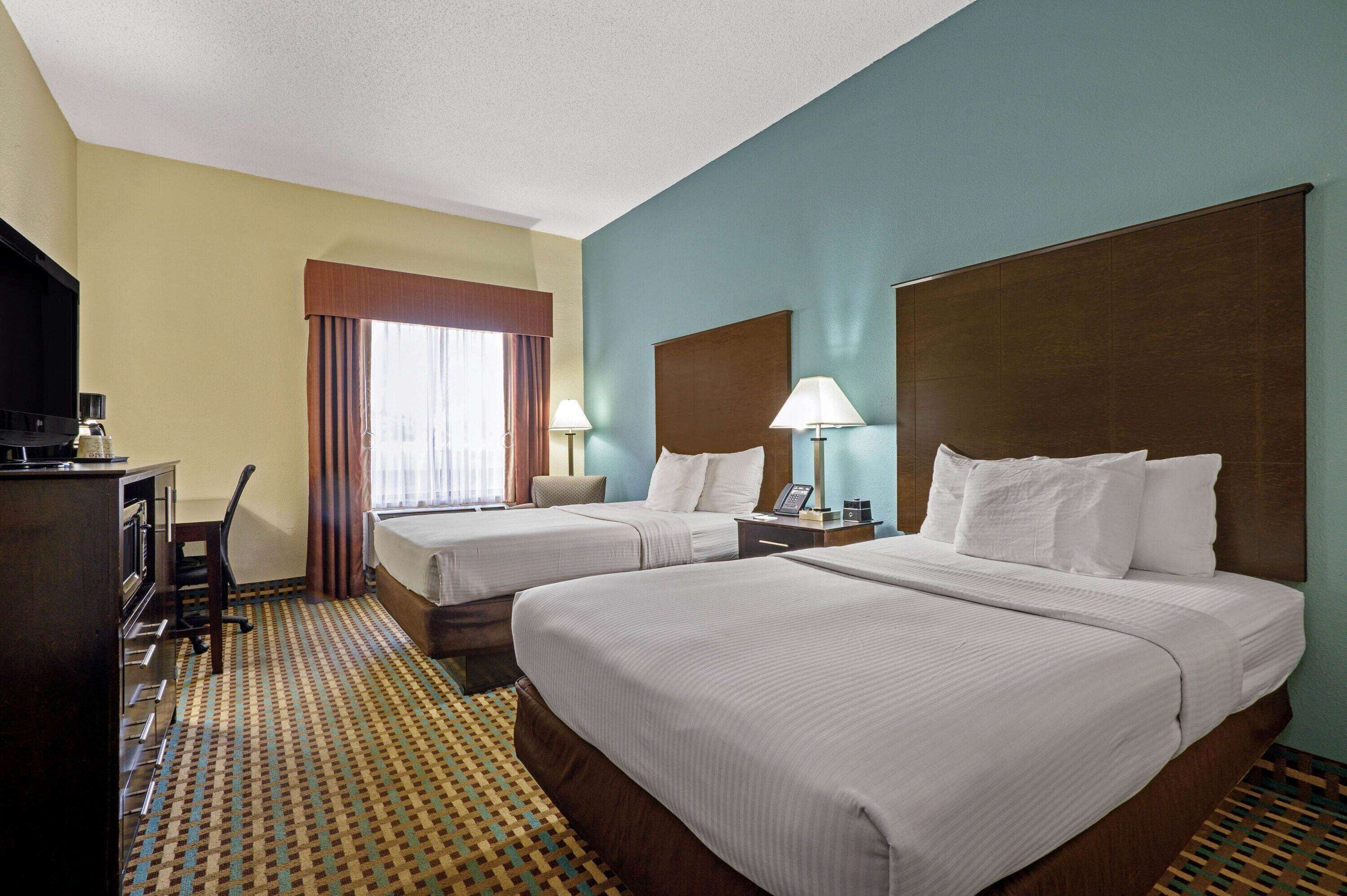 A hotel room with two beds, a desk with a lamp, a television, and a patterned carpet.