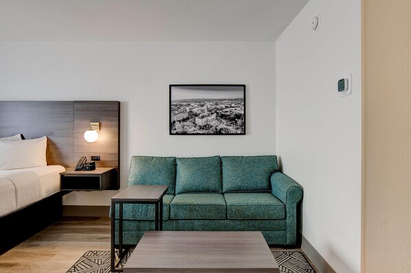 A hotel room with a bed, a green sofa, a small table, a nightstand with a lamp, and a framed cityscape picture on the wall.
