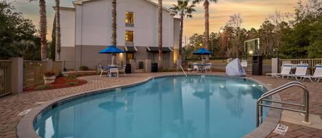 A swimming pool with lounge chairs and umbrellas, surrounded by palm trees and a building.