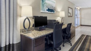 Een hotelkamer met een bureau, computer en twee stoelen. Op de muur hangt een schilderij en er is een raam met gordijnen.