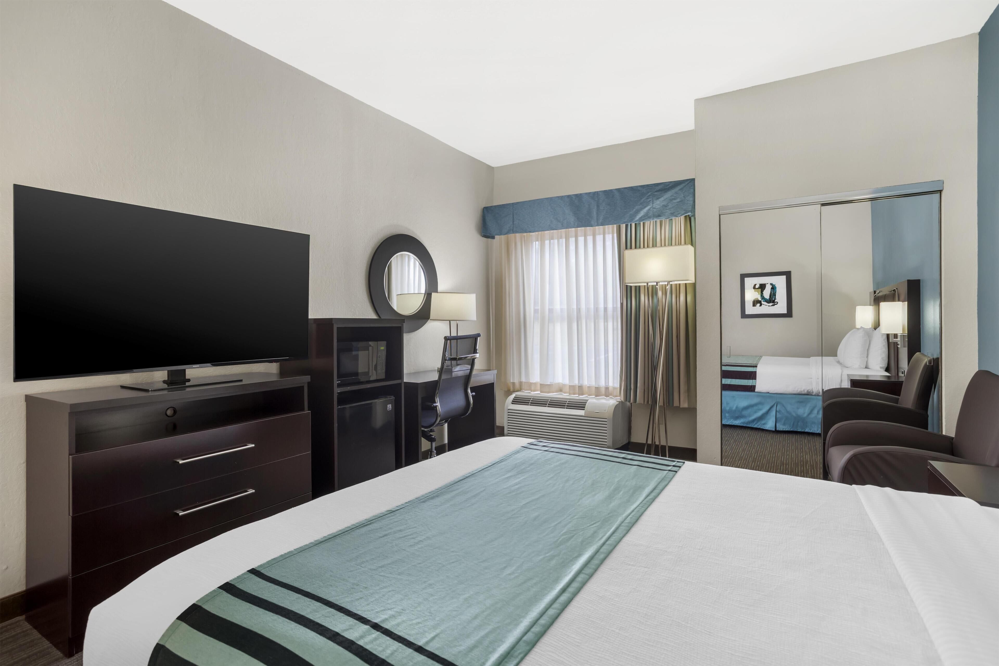 Habitación de hotel con una cama grande, un televisor de pantalla plana, un escritorio con una silla, un espejo y una ventana con cortinas.