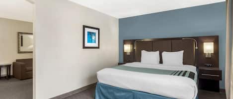 Una habitación de hotel con una cama grande, una mesita de noche con lámpara, un cuadro enmarcado en la pared y una pequeña mesa con una silla.