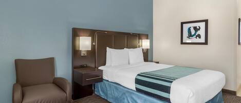 Hotellihuone, jossa on sänky, tuoli, yöpöytä ja seinään kiinnitetty valaisin.