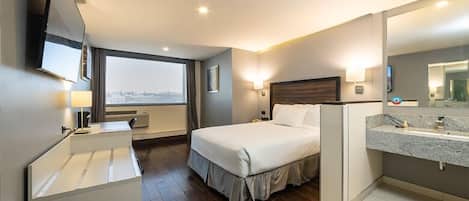 A hotel room with a large bed, a desk, a vanity with a mirror, and a window with a view of the city.