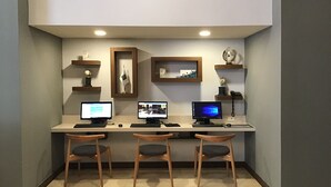 A workspace with three desks, each with a computer monitor, keyboard, and mouse. There are two wooden chairs in front of the desks. The room has a light-colored floor and walls, with shelves on the wall holding decorative items and a framed picture. - Hotel Chihuahua San Francisco (Chihuahua)