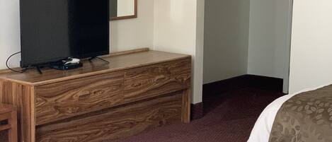 A hotel room with a bed, a wooden dresser, a television, a mirror, and a coat rack.