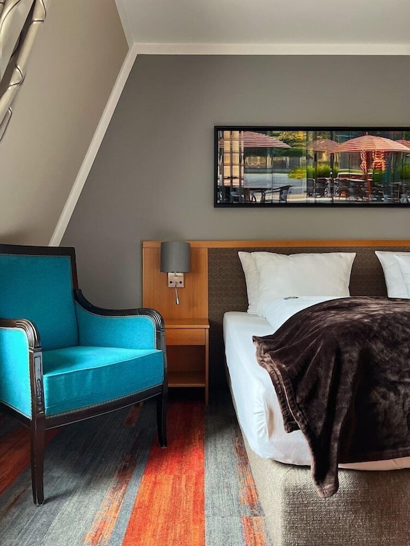 A hotel room with a bed, a blue armchair, a nightstand with a lamp, and a painting on the wall. - Prinz (Munich)
