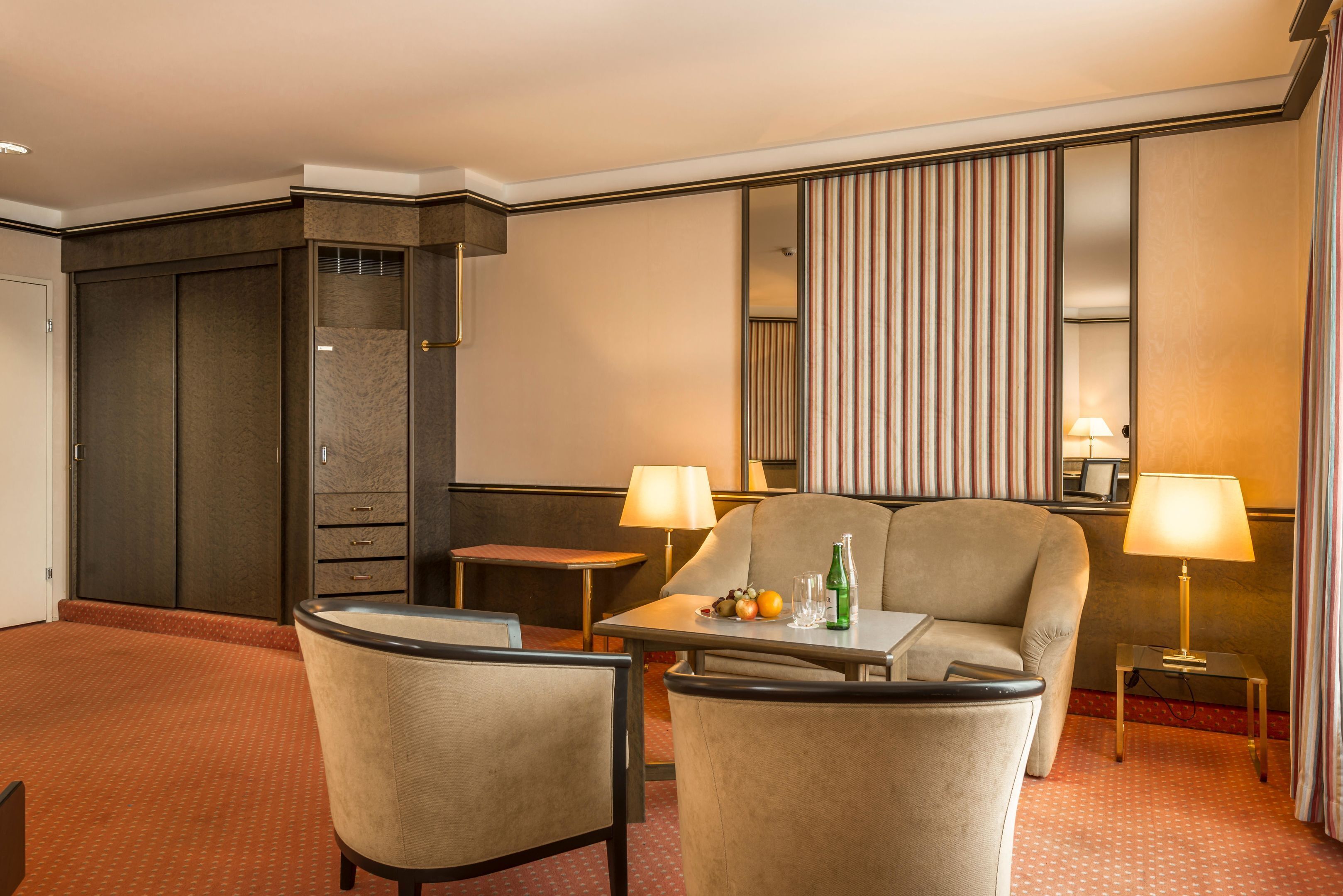 Habitación de hotel con sofá, sillón, mesita, lámparas y minibar.