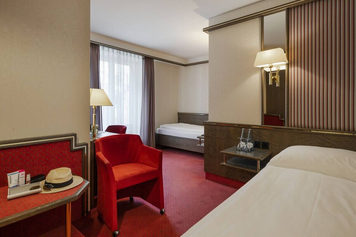 Hotellihuone, jossa on sänky, työpöytä tuolilla, punainen nojatuoli, yöpöytä lampun kanssa ja suuri peili.