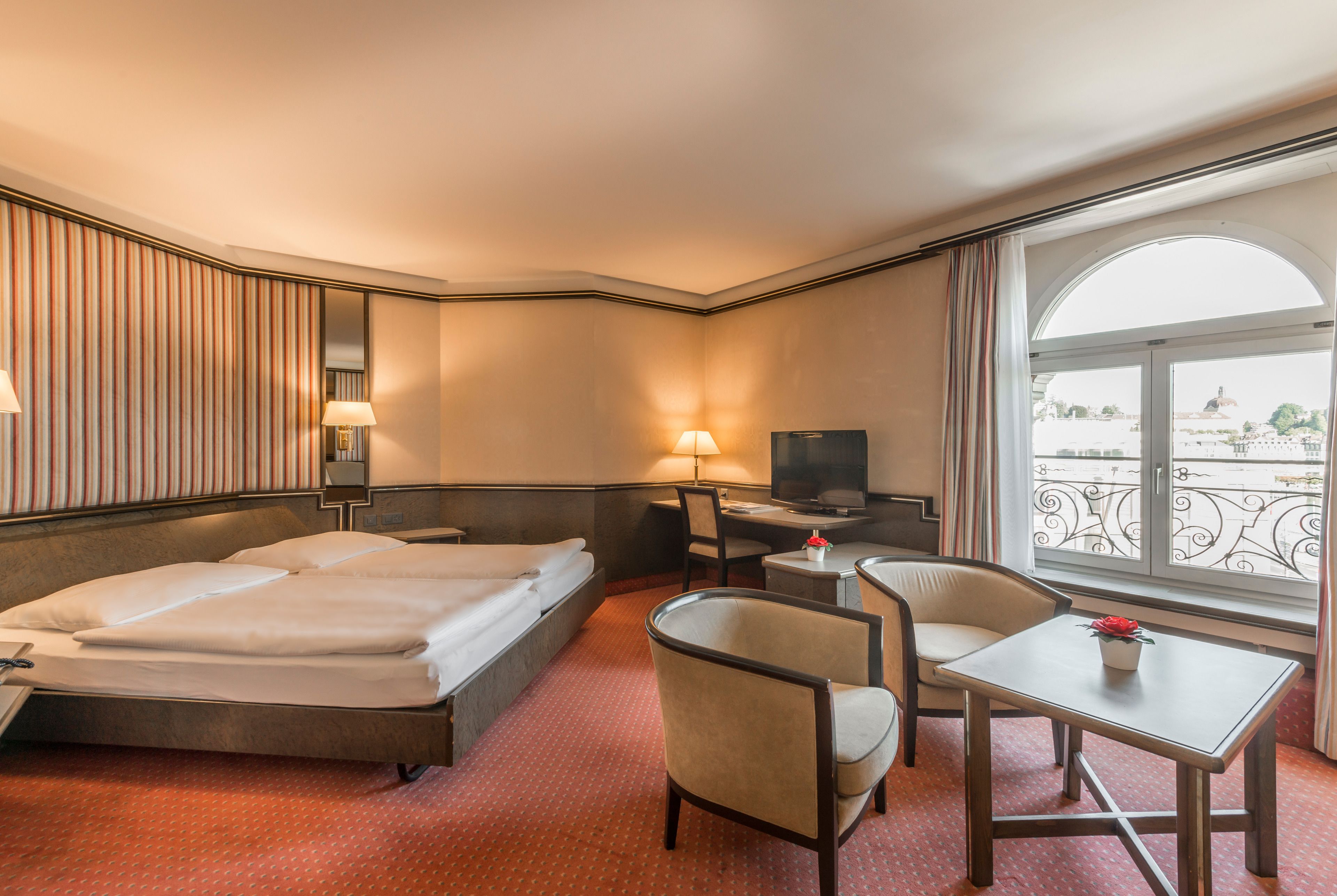 Habitación de hotel con cama, dos sillas, una mesita, televisor y ventana con vista a un edificio.